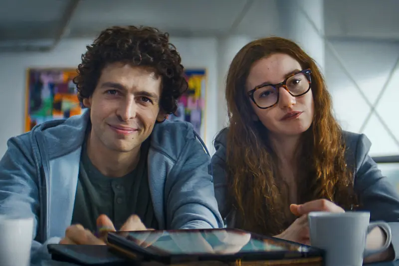 Sam Bankman-Fried (played by Anthony Boyle) and Caroline Ellison (played by Julia Garner) sitting together at a table.