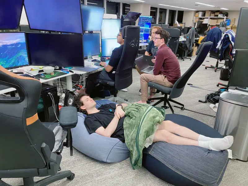 Sam Bankman-Fried sleeps on bean bag chairs in the FTX Hong Kong office while employees work at their desks in the background.